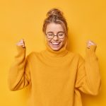 Happy positive woman with hair gathered in bun clenches fists rejoices success celebrates victory or great result wears casual jumper round spectacles poses against yellow wall. Yes I finally did it